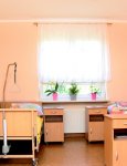 Wnętrze domu pomocy. Pokój mieszkańców. Dwa łóżka rehabilitacyjne stojące po przeciwległych stronach pokoju. Przy łóżkach szafki nocne. Kremowe ściany. W głębi okno z ażurową firaną. Na parapecie okna kwiaty doniczkowe